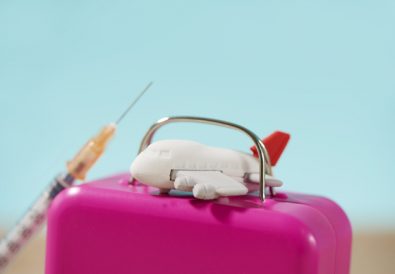 closeup of an airplane on a pink suitcase, and a syringe on the sand, on a blue background, depicting the medical tourism business