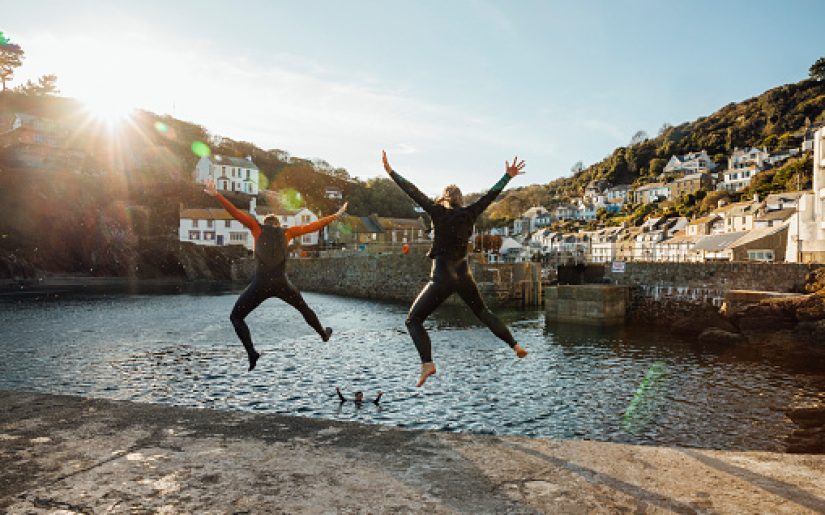 A shot of a mature woman and senior man jumping into a harbour together in wetsuits at Polperro, Cornwall.