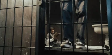 Inmate reading a book inside a prison cell while another stands nearby