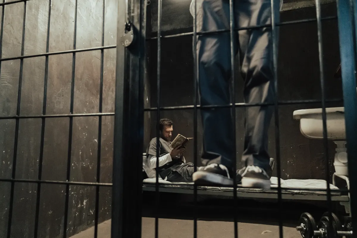Inmate reading a book inside a prison cell while another stands nearby